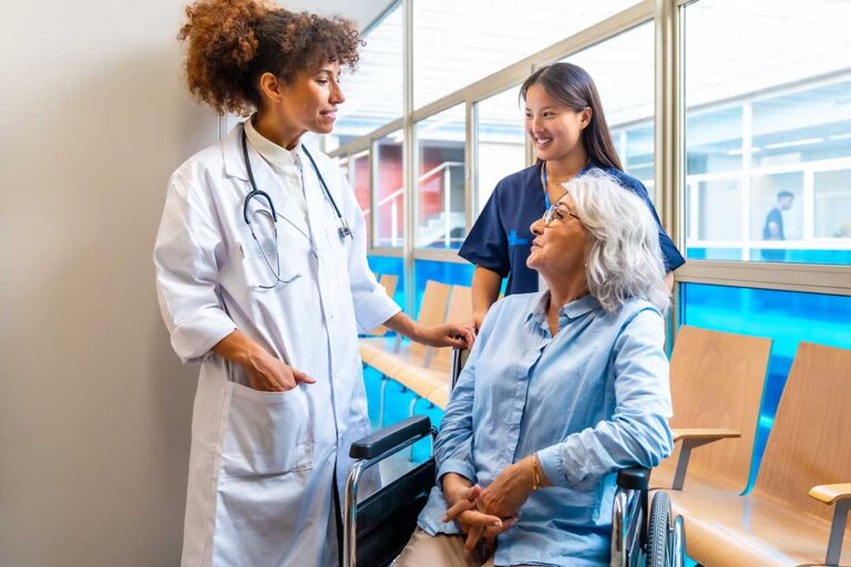 A caregiver gently assists an older adult with walking down a hallway using a walker.